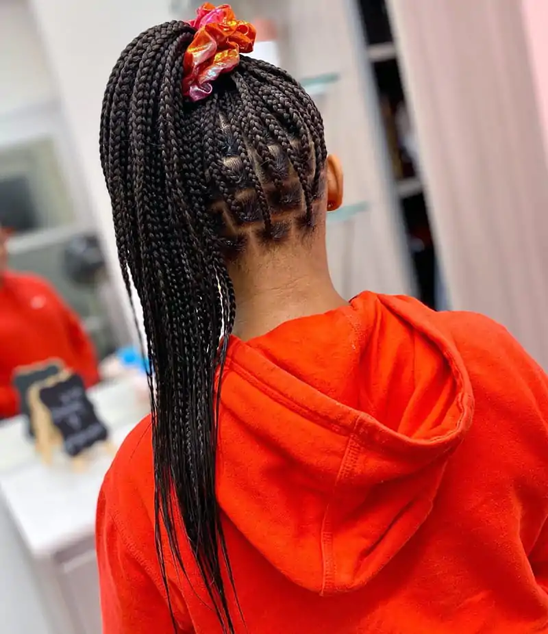 A 12 Year Old Kid wearing Knotless Box Braids. Her braids are pulled up into a pinytail and tied with a colorful hair bow